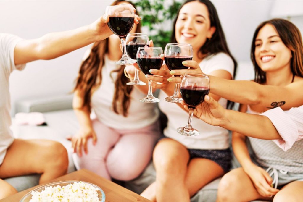 Amies jouant au jeu âJe nâai jamaisâ lors dâun EVJF dans un intĂ©rieur cosy, entre rires, cocktails et rĂ©vĂ©lations complices autour de la mariĂ©e verre de vin rouge