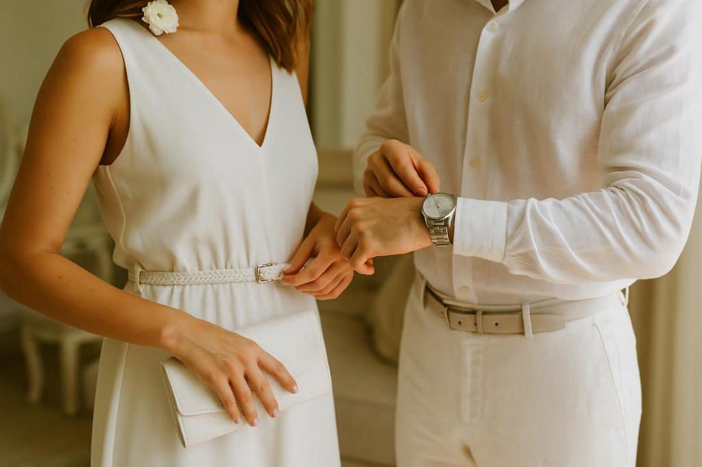 Détail d’un couple ajustant leurs tenues blanches avant une soirée chic : robe ceinturée, montre argentée, sac à main blanc et fleur dans les cheveux
