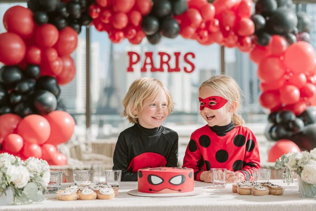 Enfants déguisés en Ladybug et Chat Noir fêtant un anniversaire dans une salle décorée rouge à pois noirs avec décor de Paris et Tour Eiffel en arrière-plan