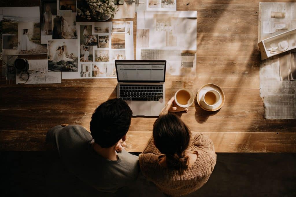 Couple préparant leur mariage en petit comité à la maison, avec ordinateur, tableau d'inspiration et budget sur table en bois, ambiance chaleureuse
