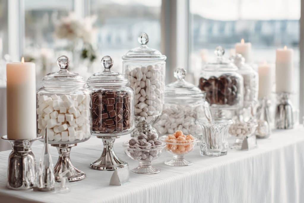 Candy bar mariage chic et minimaliste sur table blanche avec bocaux en verre et présentoirs chromés